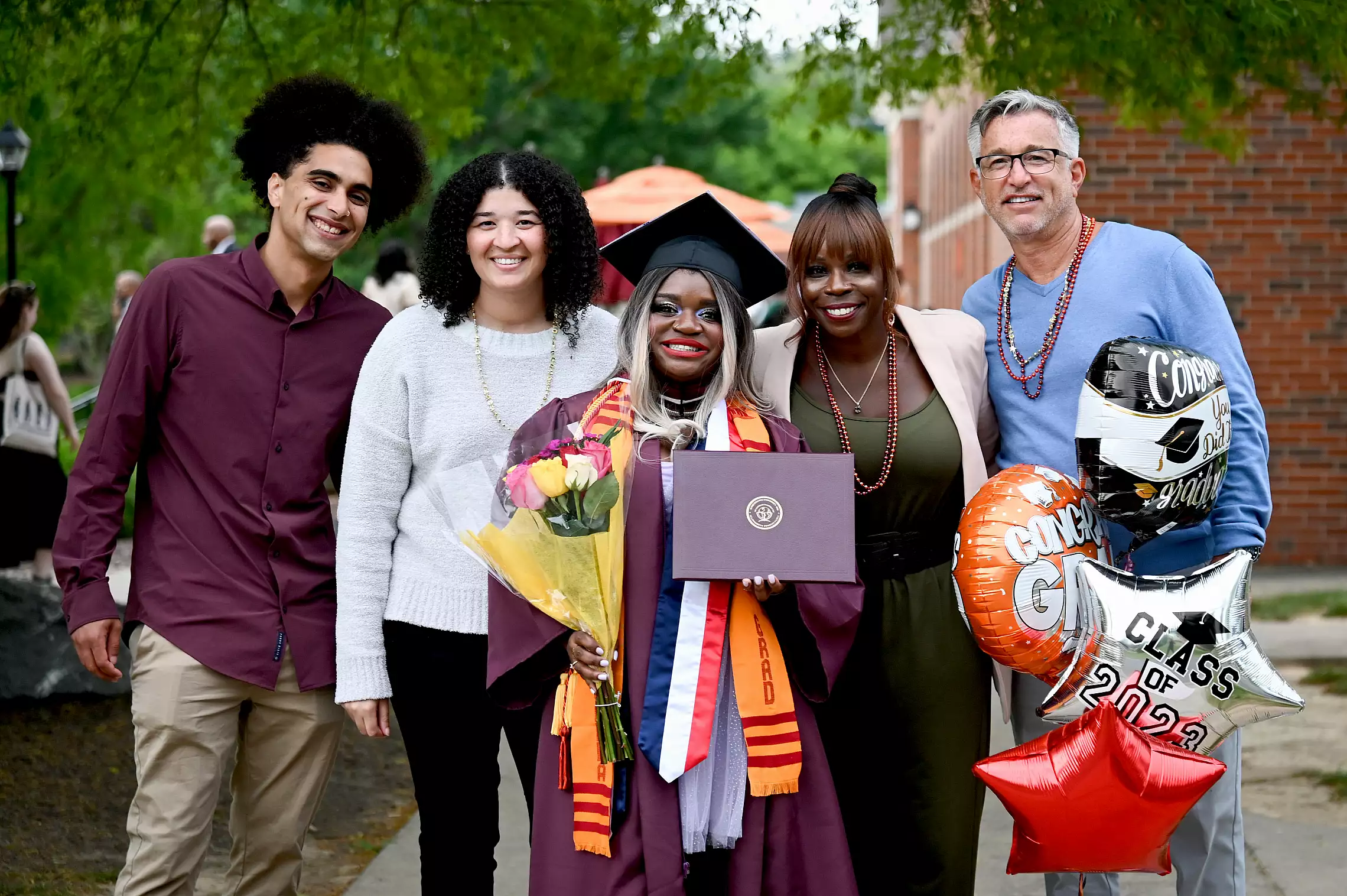 A smiling graduate in cap and gown holds flowers and a diploma at commencement, surrounded by four happy loved ones. They stand outdoors with 