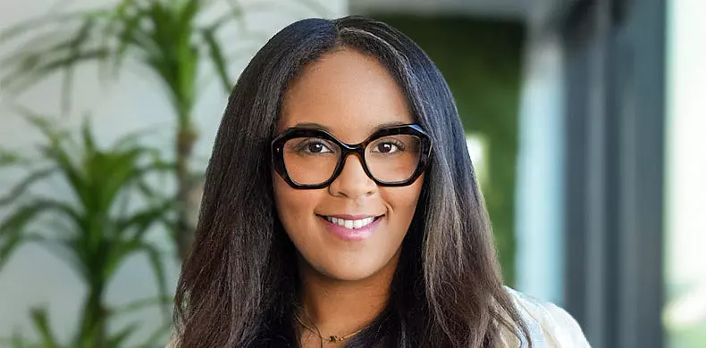 A woman with long dark hair, wearing large black glasses and a white top, smiles at the camera. There are green plants and blurred office elements in the background.