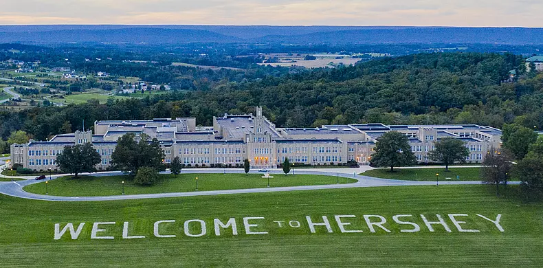 A large building sits among green lawns with trees and hills in the background. On the grass in front, white letters spell out “WELCOME TO HERSHEY.”.