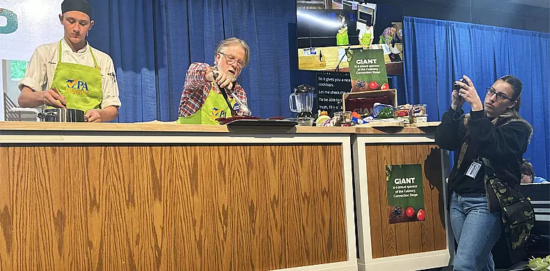 Two men at a cooking demonstration stand behind a wooden counter; one cooks while the other speaks. A woman on the right takes a photo with her phone. Vegetables and kitchen tools are displayed on the counter. Blue curtains hang behind them.