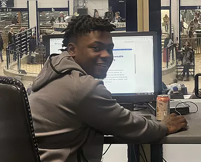 A person in a gray hoodie smiles at the camera while sitting at a desk with a computer monitor and a can of drink, with a livestock show visible through large windows behind them.