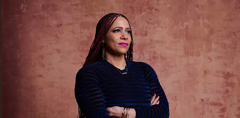 A woman with long braided hair and hoop earrings stands with her arms crossed, looking slightly upward. She wears a dark striped top and bright lipstick, with a textured brownish-pink background behind her.