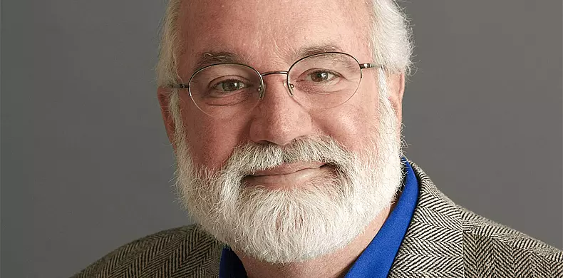 An older man with glasses, a white beard, and white hair, wearing a blue shirt and a gray plaid blazer, smiles slightly against a plain gray background.