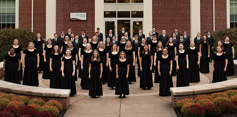 A large choir group poses outside a brick building. The women wear long black dresses and the men wear black suits. They stand in rows on a curved walkway, smiling at the camera, with flowers and greenery in the foreground.
