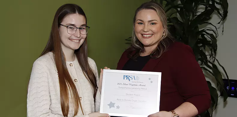 Two women smiling; one wearing glasses and a cream sweater, the other in a maroon blazer. They are holding a certificate together with a PRSA logo, standing in front of a green wall and some plants.