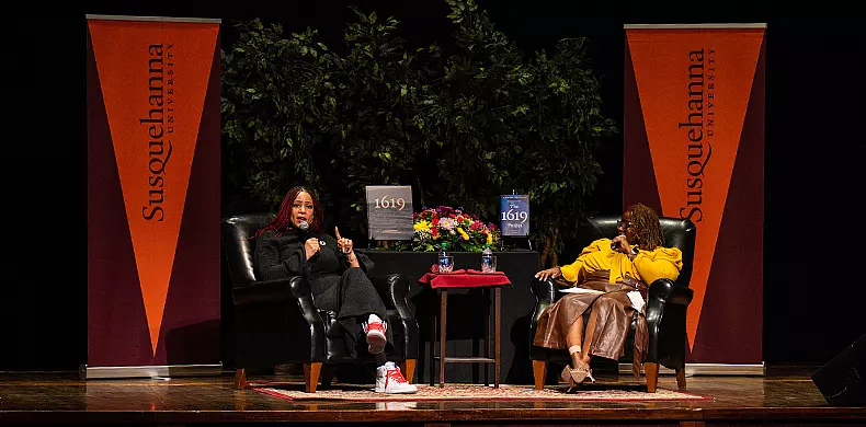 Two women sit on stage in armchairs between two orange 体育买球 University banners, speaking at an event. A table with flowers and books is between them and green plants are in the background.