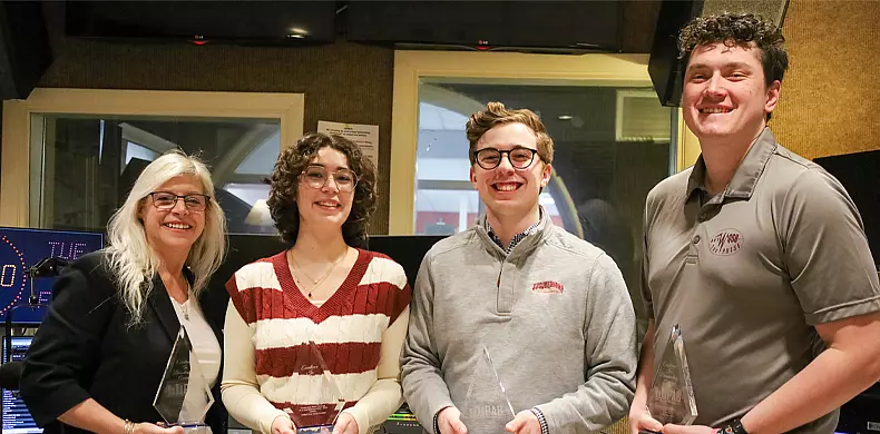 Four people stand indoors, smiling and holding clear awards. One woman is wearing glasses and a white jacket, the others wear casual tops. They appear to be in a recording or broadcast studio.