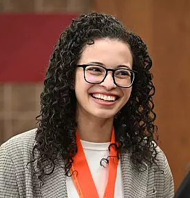 A woman with curly hair and glasses smiles warmly. She is wearing a white top, a gray blazer, and an orange lanyard around her neck. The background is softly blurred with warm tones.