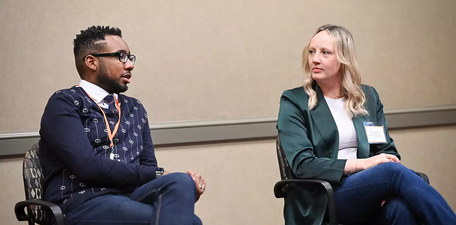 Two people sit in chairs facing each other, engaged in conversation. The man on the left wears glasses, a patterned sweater, and tie. The woman on the right has blonde hair, wears a green blazer, and has a conference badge.