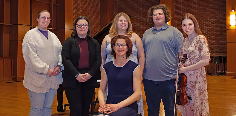 Six people pose indoors on a wooden stage with a grand piano and brick wall in the background. Five are standing, one is seated in front; one person holds a violin. All are smiling and dressed in casual or semi-formal attire.