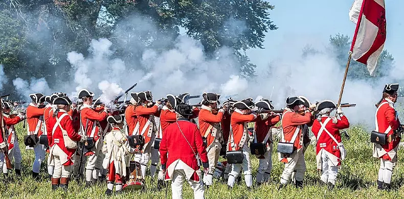Historical reenactors dressed as British Redcoat soldiers in red uniforms and black hats fire muskets in formation on a grassy field, with smoke rising and trees in the background.