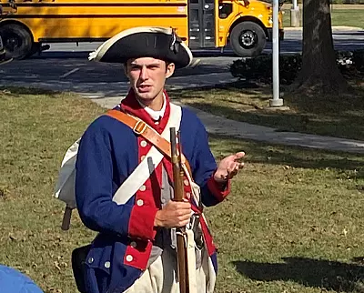 A person dressed in a Revolutionary War-era soldier costume holds a musket and gestures while standing outside; a yellow school bus is visible in the background.