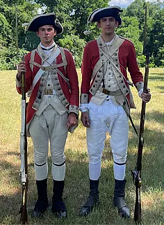 Two men stand side by side outdoors in 18th-century British redcoat military uniforms, holding long rifles and wearing black hats, white breeches, and tall black boots, with trees and grass in the background.