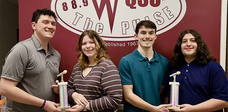 Four smiling young adults stand in front of a maroon and white QSG Pulse radio sign. Two of them hold gold microphone trophies, and all are casually dressed, posing together for a group photo.