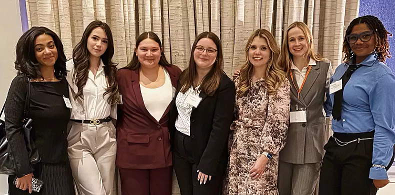 Seven women are standing side by side indoors, smiling at the camera. They are dressed in business or semi-formal attire, with a curtain in the background. Some are wearing name badges.