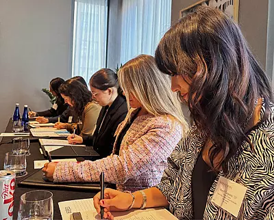 A row of women sit at a conference table, focused on taking notes during a meeting. Notepads, glasses, bottled water, and a can of Diet Coke are on the table. Bright windows provide natural light in the room.