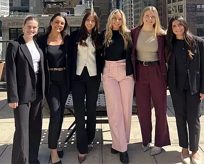Six women stand side by side on a rooftop, smiling at the camera. They are dressed in business or business-casual attire, with tall buildings visible in the background.