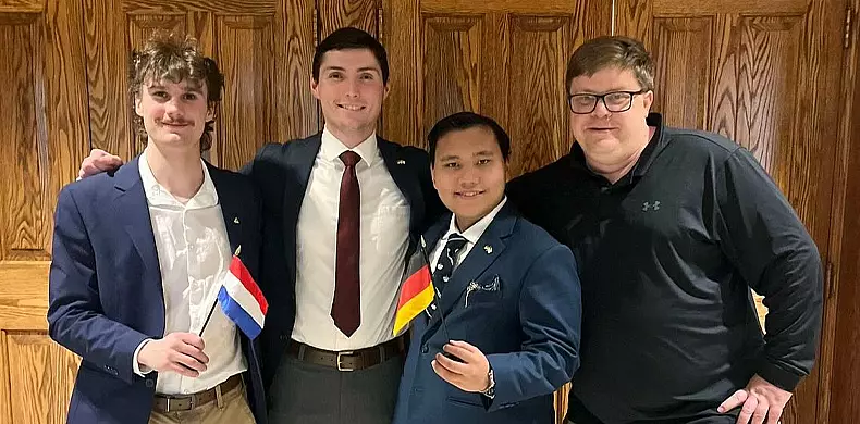 Four men stand smiling in front of a wooden wall. Two of them are holding small flags: one holds a Russian flag and the other holds a German flag. They are dressed in suits and casual attire, posing together.