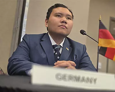 A person in a suit sits at a table with a microphone, a small German flag, and a nameplate labeled 
