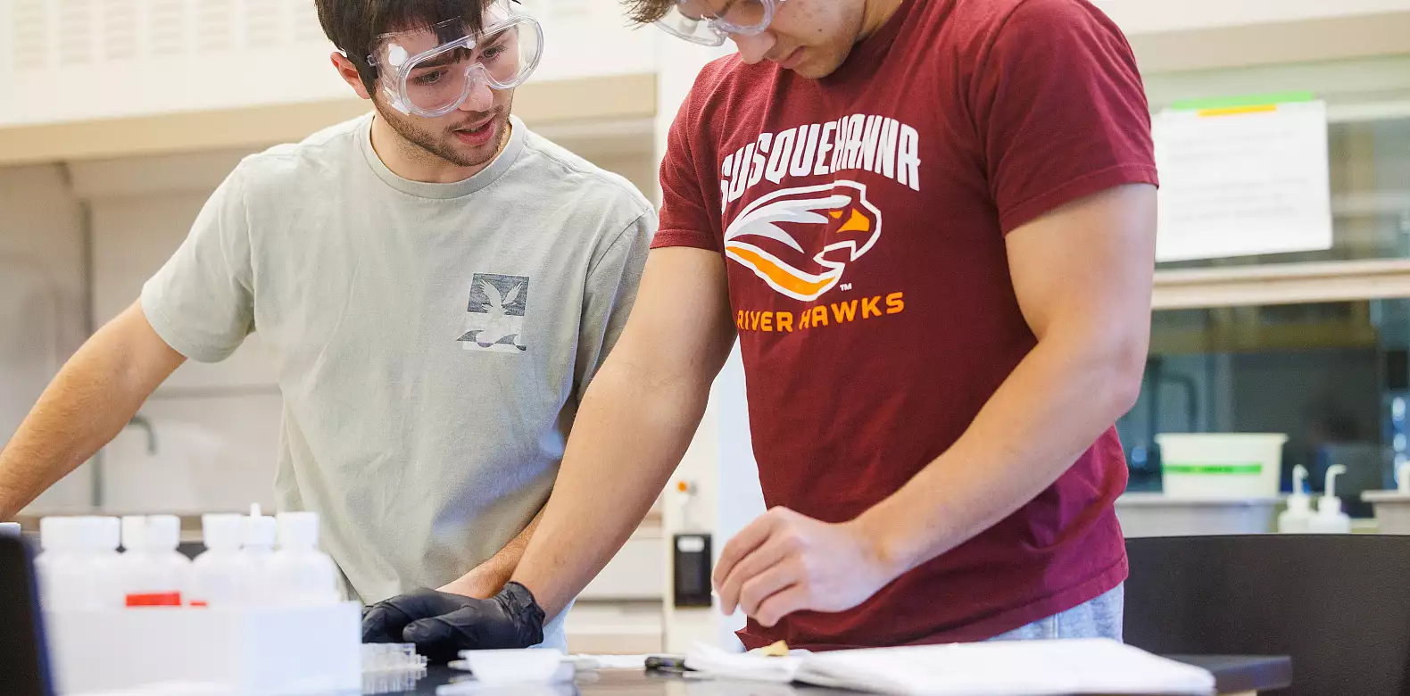 Two students wearing safety goggles work together in a science lab, examining materials on a lab bench with bottles and tools in front of them. One student wears gloves; the other wears a River Hawks t-shirt.