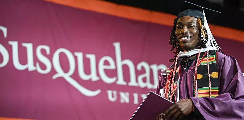 A smiling graduate in a cap and gown holds a diploma in front of a maroon 体育买球 University banner.