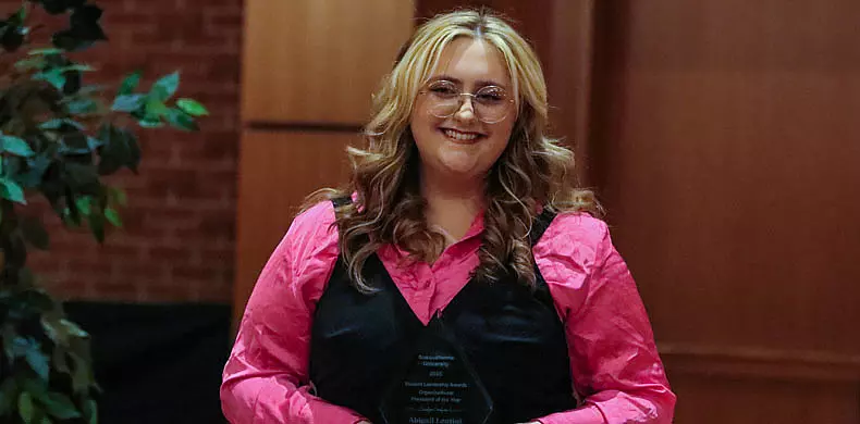 A person with long blonde hair, glasses, and a bright pink shirt under a black vest smiles while holding an award in a formal indoor setting.
