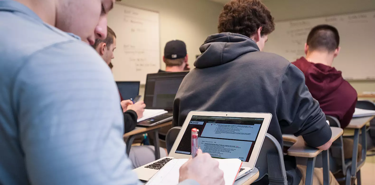 Students sitting at desks in a classroom, some using laptops and notebooks to take notes, with whiteboards and notes visible in the background.