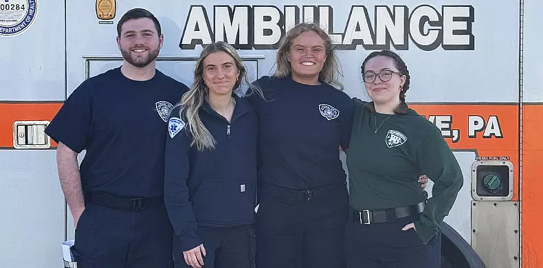 Four smiling people in uniform stand arm-in-arm in front of an ambulance. Three wear navy shirts, one wears a green shirt. The ambulance has 