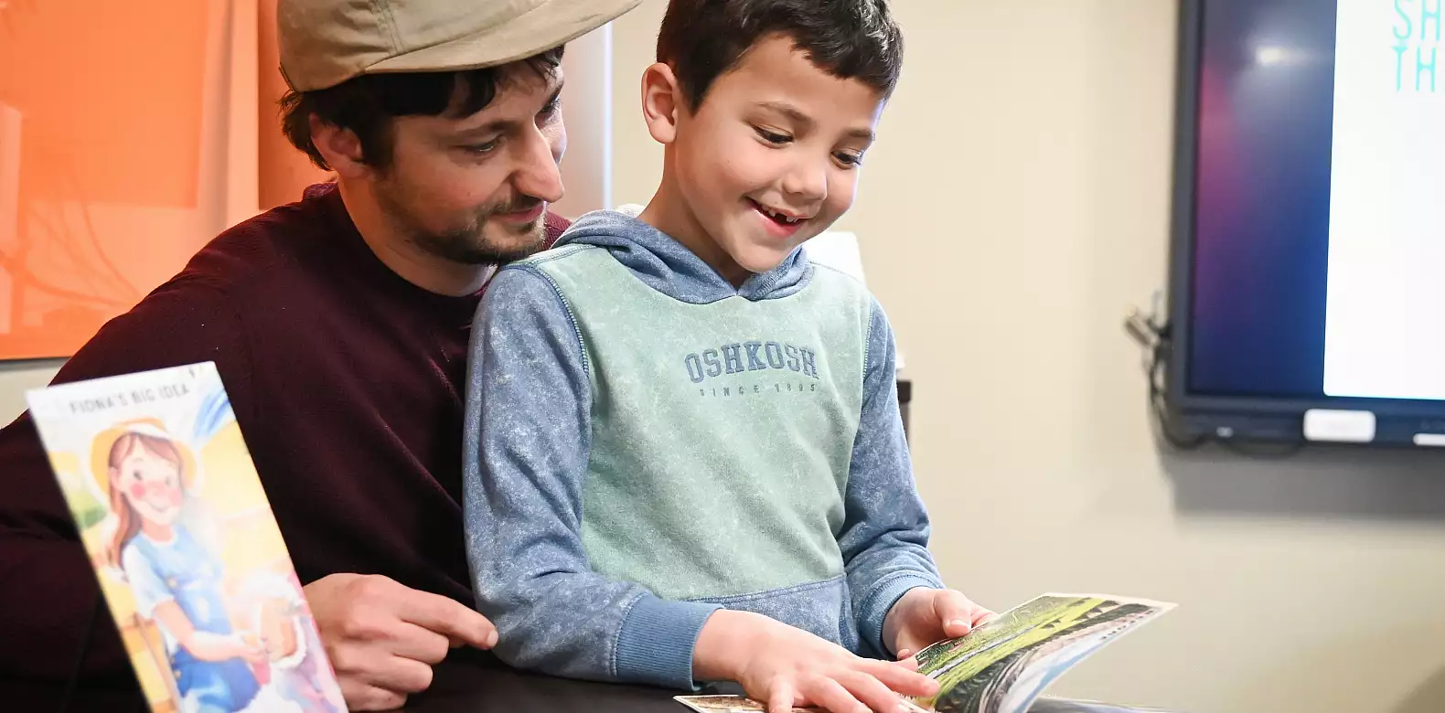 An adult and a smiling young boy sit together at a table, reading a book. Another illustrated book stands upright nearby. The adult looks at the boy, while the boy points at the open book’s pages.