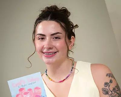 A smiling young woman with dark hair in a bun, wearing a sleeveless light top, colorful beaded necklace, and braces, holds up a book. She has a floral tattoo on her upper arm.