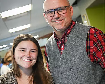 A smiling young woman with long brown hair stands next to a man wearing glasses, a gray vest, and a red plaid shirt. They are indoors, likely in an office or classroom setting.