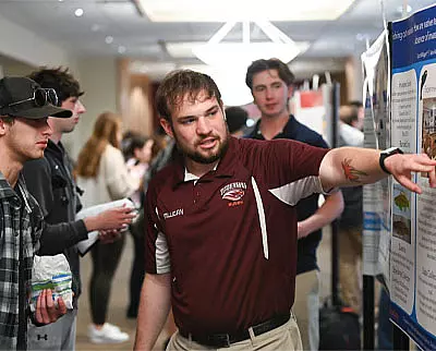 A man in a maroon shirt points to a poster board while explaining something to a group of people at an indoor event. Others look on attentively in a busy, well-lit room.