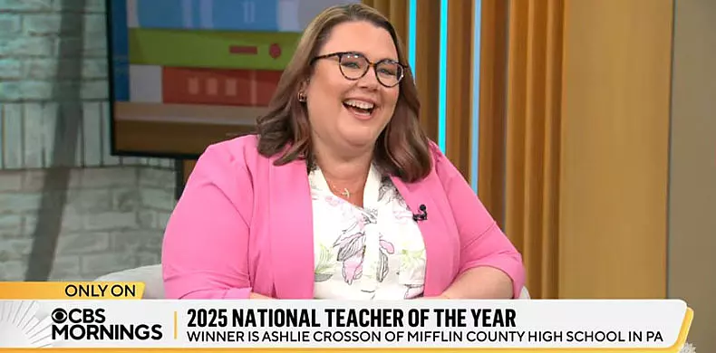 A woman wearing glasses and a pink blazer smiles while seated on a TV set. The screen text reads: 