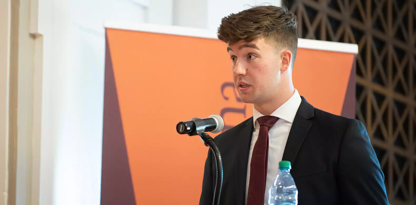 A young man in a suit and tie speaks into a microphone at a podium, with a bottle of water in front of him and an orange banner in the background.