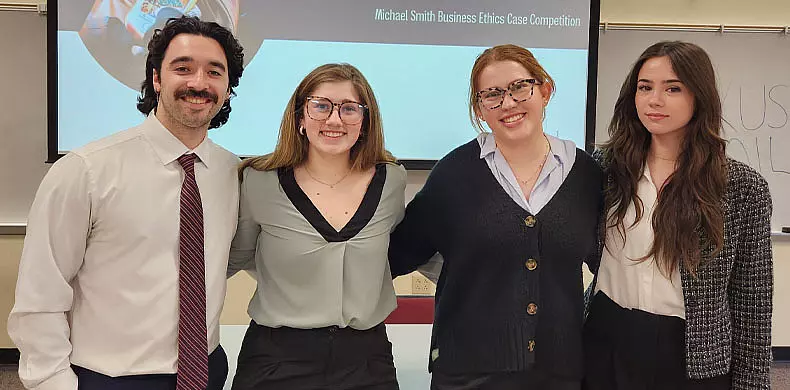 Four young adults in business attire stand smiling together in a classroom, with a presentation slide behind them reading “Michael Smith Business Ethics Case Competition.”.
