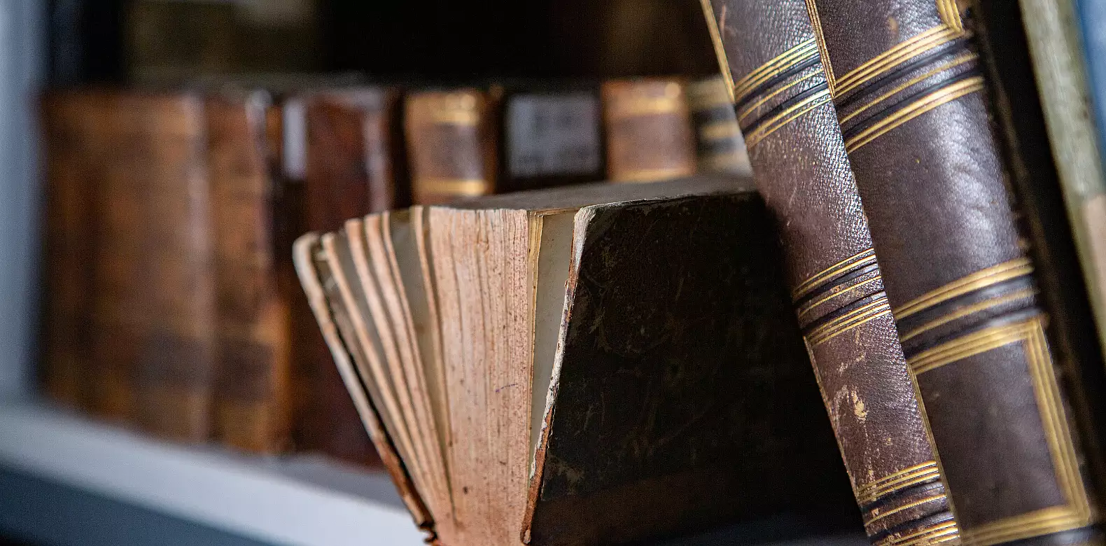 A close-up of several old, worn books with faded pages and brown, textured covers sitting on a bookshelf. The spines and edges show signs of age, with some books stacked upright and others leaning.