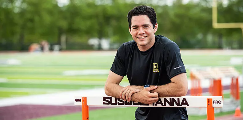 A man in a black Army t-shirt smiles while leaning on a hurdle labeled 
