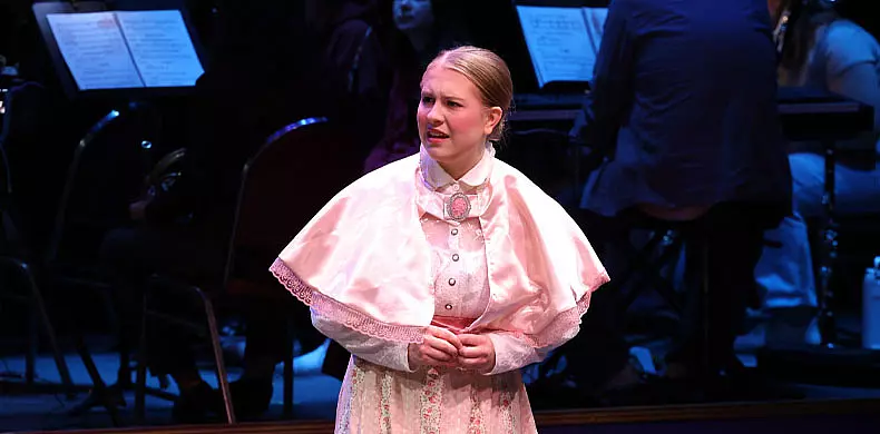 A woman in a pink vintage dress and cape performs on stage, looking concerned, with an orchestra seated and playing in the background.