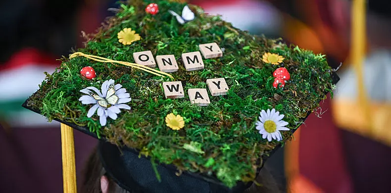 A graduation cap decorated with green moss, faux flowers, small mushrooms, and Scrabble tiles spelling “ON MY WAY.”.