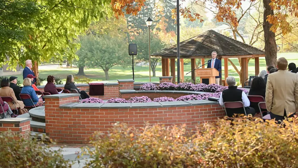 A man speaks at a podium in an outdoor amphitheater surrounded by seated people, autumn trees, and pink flowers, with a wooden gazebo in the background highlighting Ways to Give.