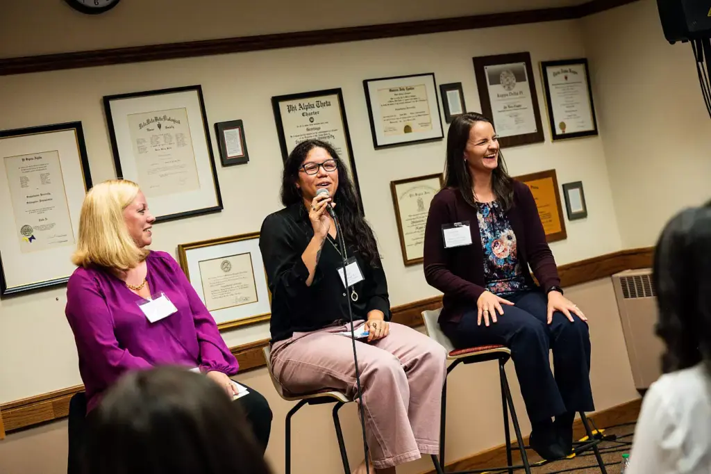 Three women sit on stools, smiling and speaking at a panel discussion about Ways to Give. One woman in the center holds a microphone. Certificates and awards are displayed on the wall behind them, with an audience partially visible in the foreground.