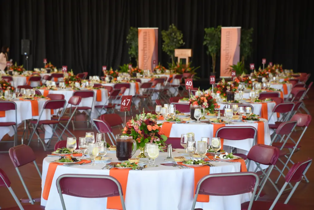 A banquet hall with round tables covered in white and orange tablecloths, set with plates, glasses, flowers, and food. Maroon chairs surround the tables; banners and greenery decorate the background.