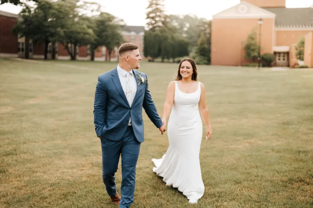 A bride in a white dress and a groom in a blue suit walk hand in hand across a grassy lawn, smiling at each other, enjoying the community access to the charming brick buildings and trees in the background.