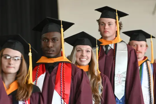 Five students in maroon graduation gowns and caps stand in a row, some smiling and wearing honor cords and stoles, during a commencement ceremony.