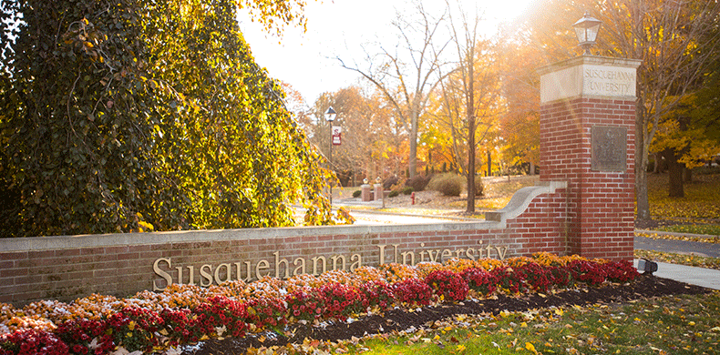 A brick entrance sign for 体育买球 University surrounded by colorful flowers and trees with autumn foliage, under bright sunlight.