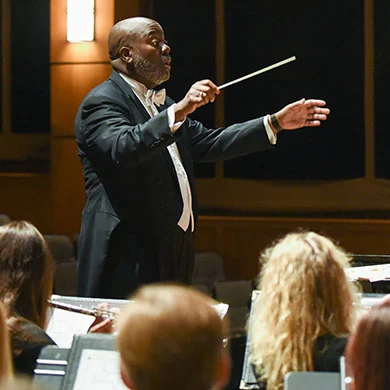 A conductor in a tuxedo leads a seated orchestra in a concert hall, passionately directing musicians as they focus on their sheet music, creating an inspiring atmosphere of community access to live music.