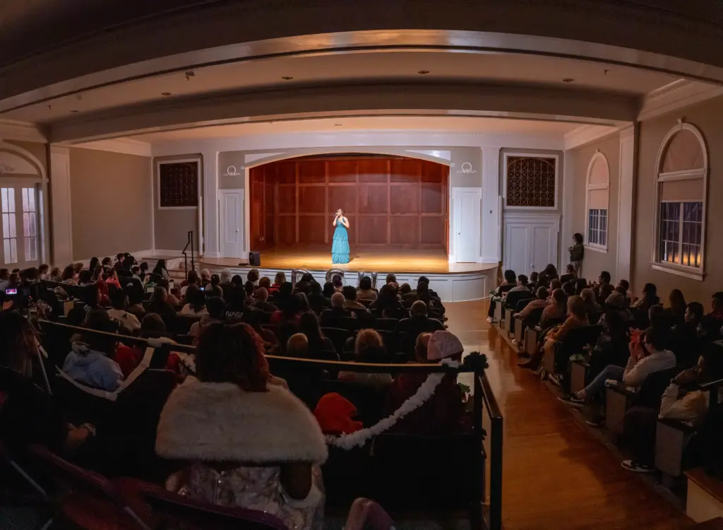 A performer in a blue dress stands on a well-lit stage, addressing a seated audience in a large, elegant auditorium with arched windows and wooden floors.