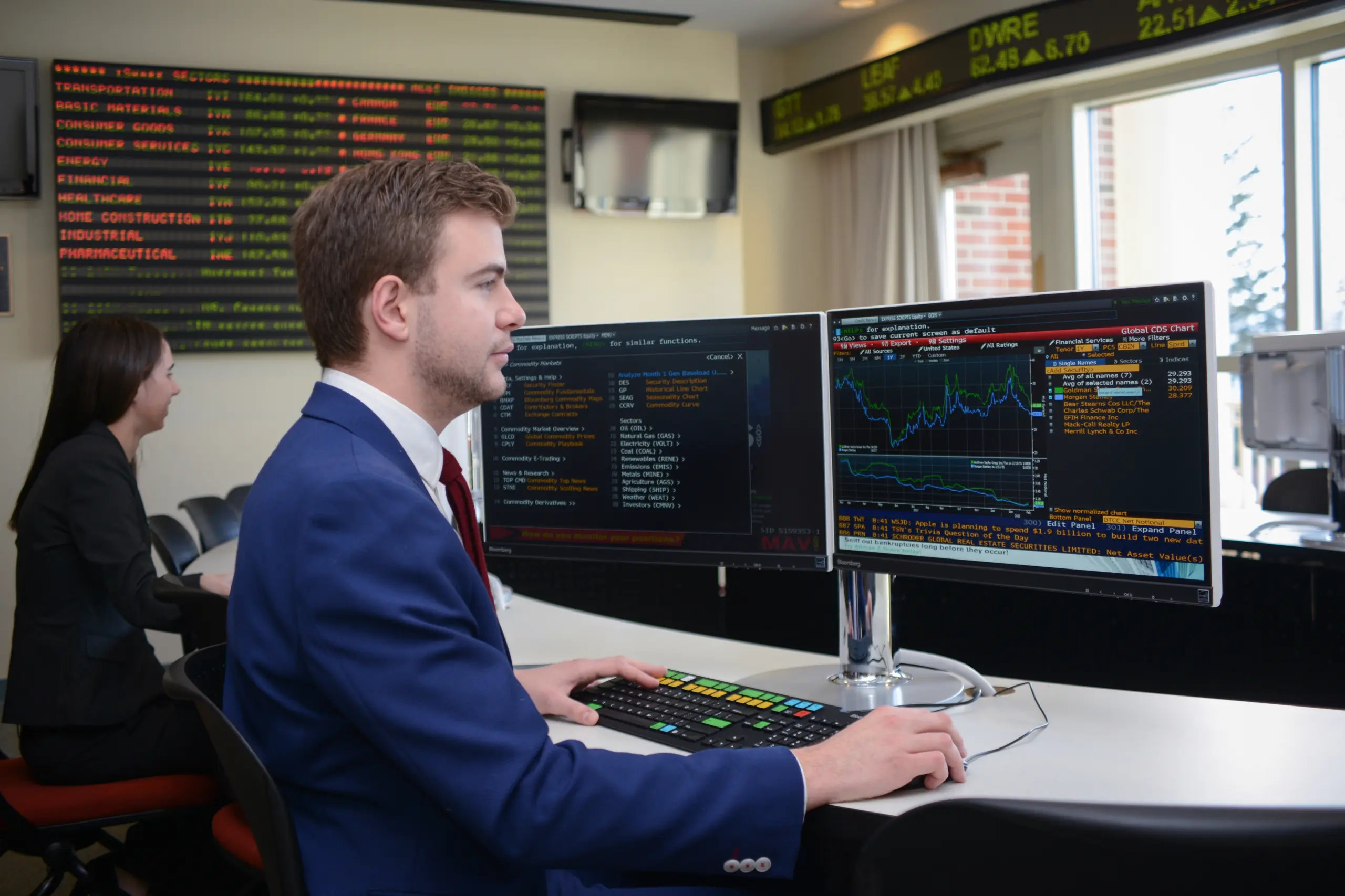 A man in a blue suit sits at a desk, analyzing financial data on large computer monitors. A woman works in the background. Stock market boards on the walls highlight a Learn By Doing approach to finance.