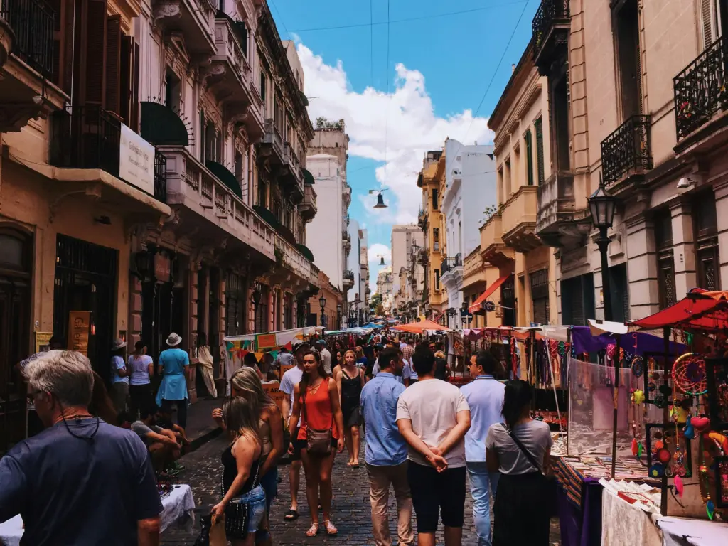 A lively outdoor market set on a narrow city street, where people stroll among colorful stalls selling various goods—an experience you might encounter when you GO Study Abroad—surrounded by old buildings under a bright blue sky with scattered clouds.