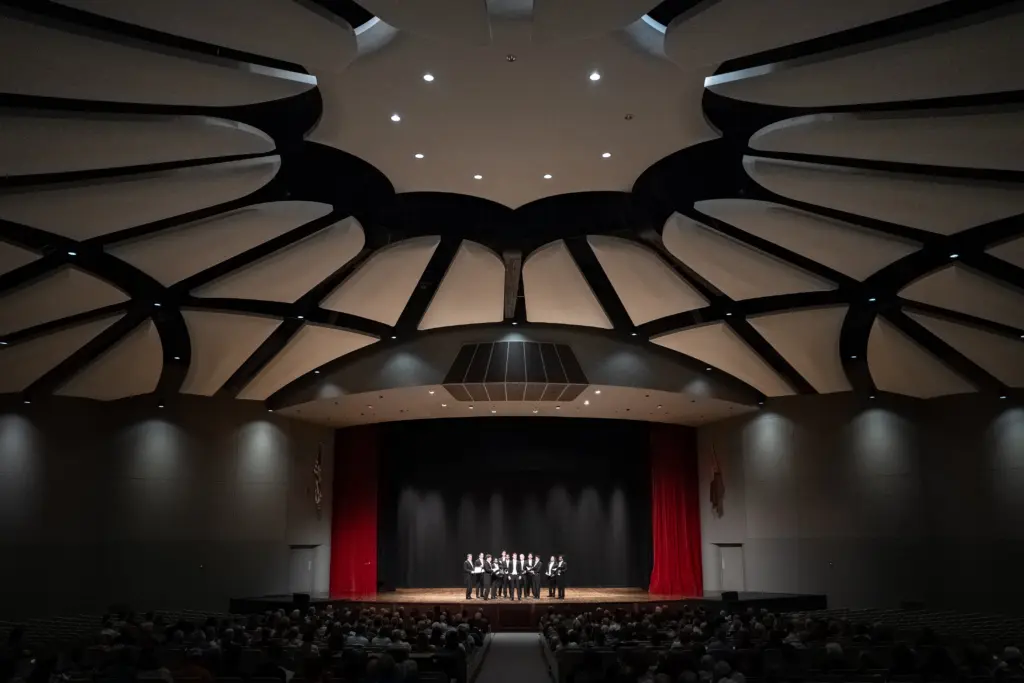 A wide-angle view of a concert hall with a dramatic, modern ceiling. A small group of performers is onstage under bright lights, while an audience enjoys the show—highlighting community access to live performances.